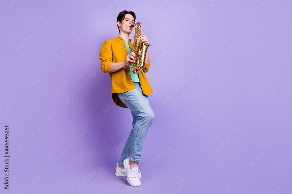 Full size photo of joyful brunet young guy play saxophone wear shirt jeans isolated on violet color background Full size photo of joyful brunet young guy play saxophone wear shirt jeans isolated on violet color background