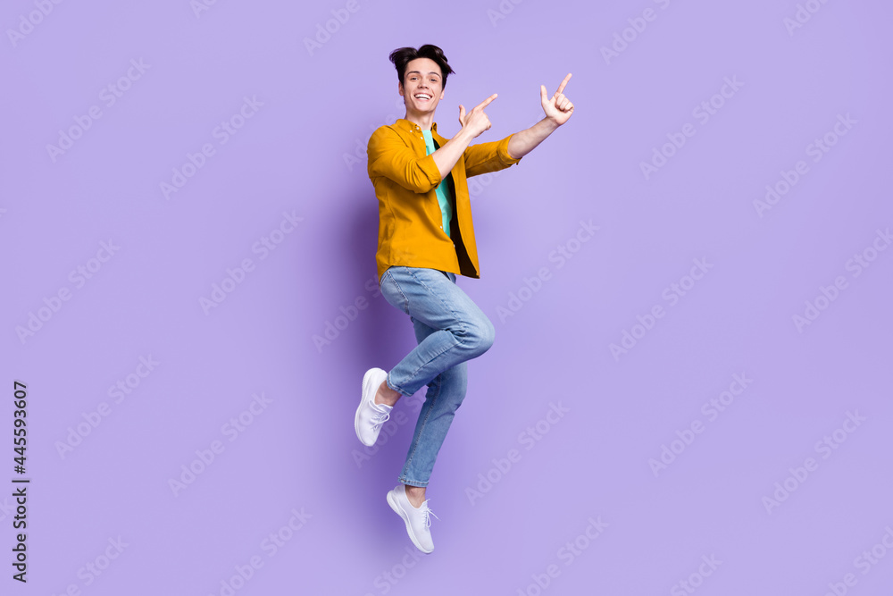 Full size photo of cool brunet millennial guy jump point empty space wear shirt jeans isolated on violet color background