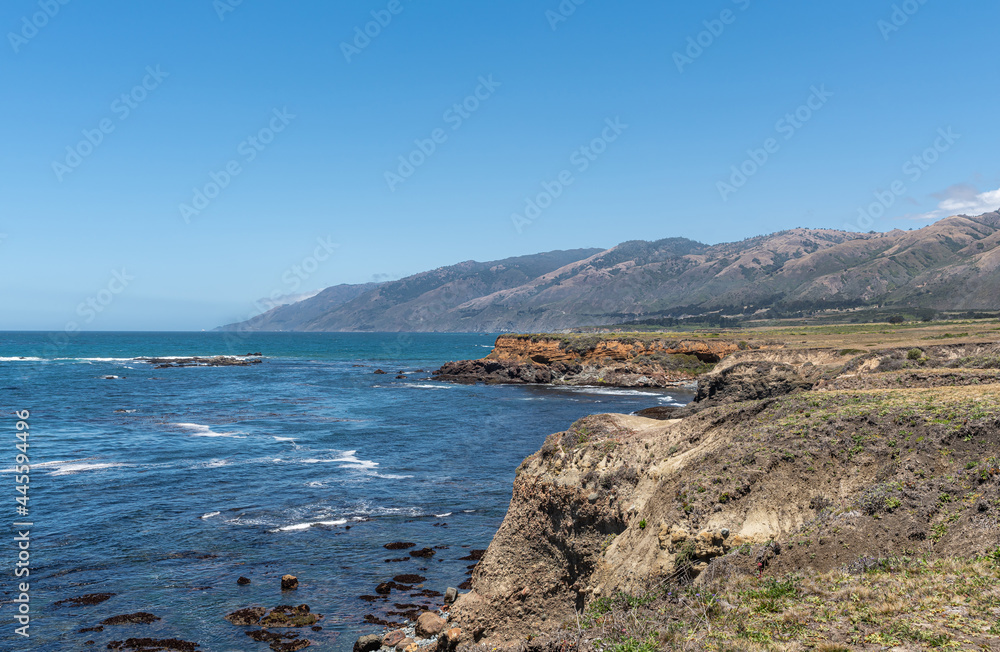 San Simeon, CA, USA - June 8, 2021: Pacific Ocean coastline north of town. Green plateau ending on red cliffs into deep blue water under light blue sky. Los Padres Mountain range.