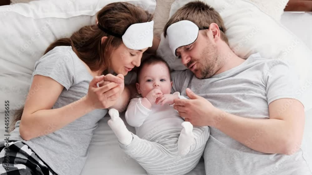 Beautiful family wearing same pajamas and sleeping eye masks lying in the bed with little child boy and petting him. Young parents with their toddler baby in bedroom. Father kisses hand of his kid Beautiful family wearing same pajamas and sleeping eye masks lying in the bed with little child boy and petting him. Young parents with their toddler baby in bedroom. Father kisses hand of his kid