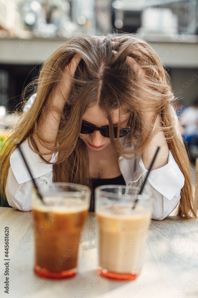 The girl cannot choose which drink to drink. Humor, funny. Ice coffee in a tall glass with milk. Old rustic wooden table. Cold summer drink with tubes restaurant. The girl cannot choose which drink to drink. Humor, funny. Ice coffee in a tall glass with milk. Old rustic wooden table. Cold summer drink with tubes restaurant.