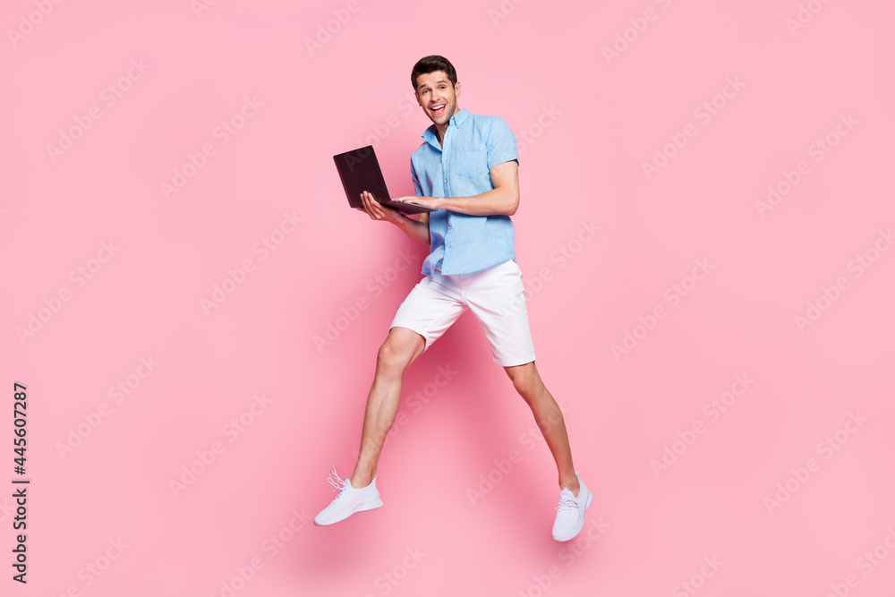 Photo of positive carefree man jump typing notebook run wear blue shirt shorts isolated on pink color background Photo of positive carefree man jump typing notebook run wear blue shirt shorts isolated on pink color background