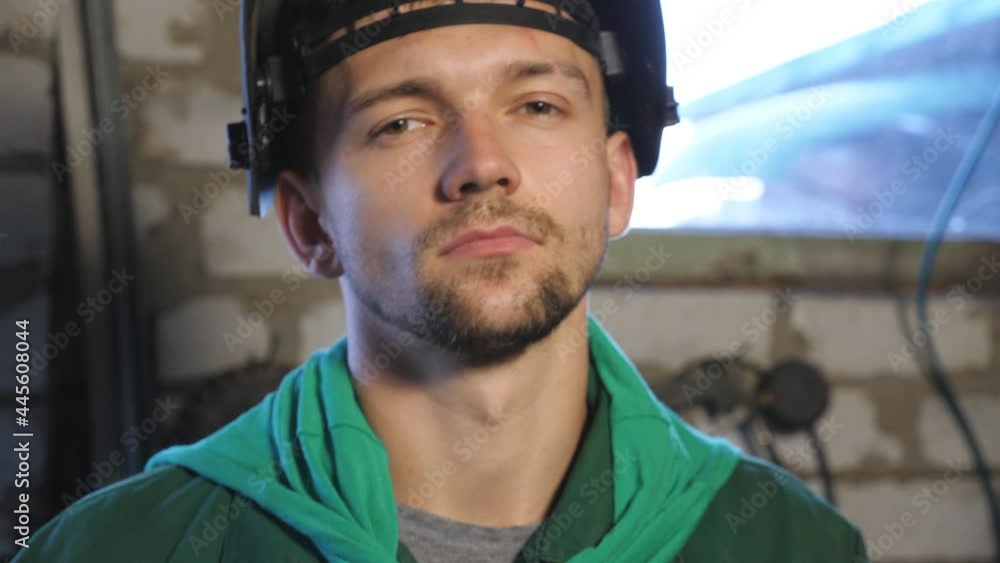 Close up of tired young repairman opens protective mask after finishing his job. Welder in workwear looking into camera in his garage. Portrait of mechanic with beard working in workshop. Dolly shot Close up of tired young repairman opens protective mask after finishing his job. Welder in workwear looking into camera in his garage. Portrait of mechanic with beard working in workshop. Dolly shot