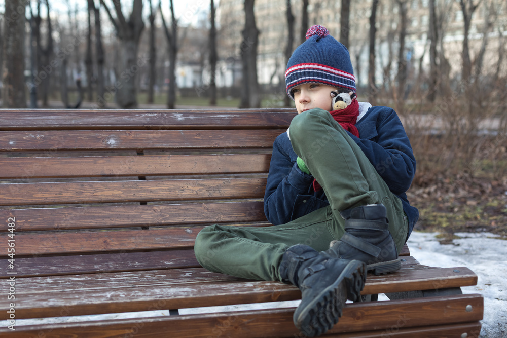 Caucasian boy on a walk in the park sits alone on a bench. Sad and sad child. The concept of the relationship problem Caucasian boy on a walk in the park sits alone on a bench. Sad and sad child. The concept of the relationship problem
