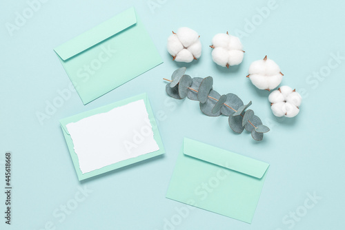Blank paper card mockup with torn edges, envelopes, cotton flowers and eucalyptus branch on a mint blue background. Wedding stationery template. Top view, flat lay.