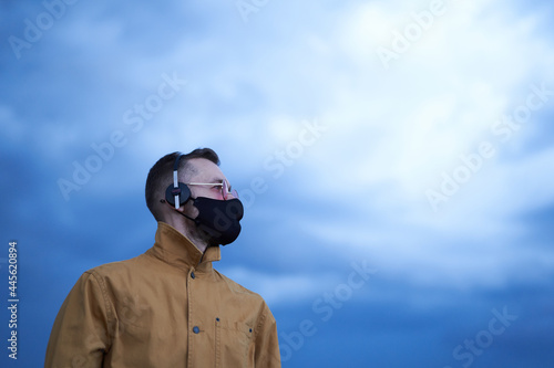 Portrait of beautiful caucasian man in pink eyeglasses, black medical face mask and orange jacket with headphones. Concept of listening to the music or soundtrack. Blue sky and rainy weather © boytsov