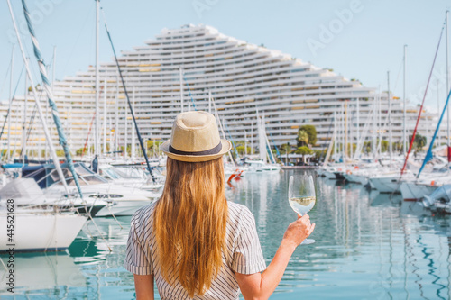 Blonde hair woman in a hat standing with her back on sea, yachts, buildings and boats background. White wine glass in hand. Vacation in Europe. Nice, French riviera. Travel photo. photo