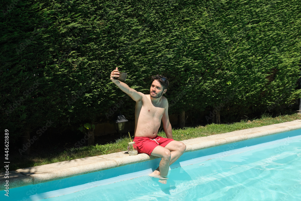 Young man taking a selfie with his smartphone sitting on the swimming pool edge. Summer concept.