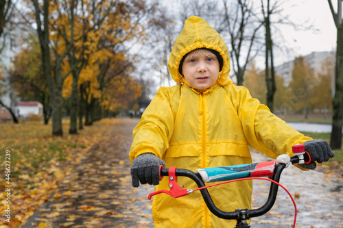 Portrait of serious preschooler in yellow raincoat. Boy rides bicycle in an autumn park in the rain.