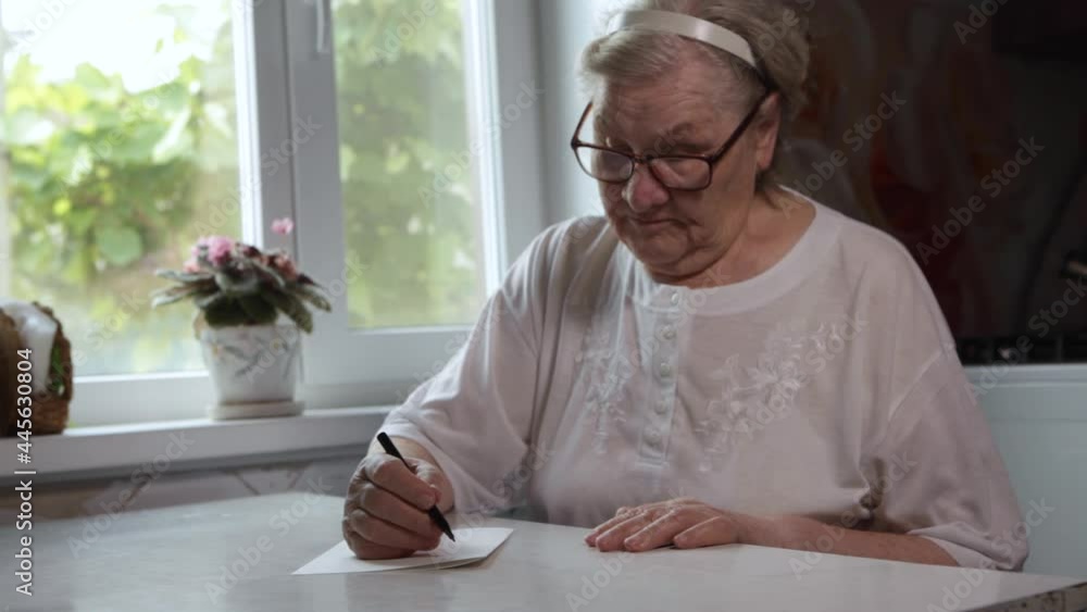 An elderly woman writes at home on a piece of paper. Hobbies of pensioners. Remote communication, sending emails. The process of writing a note An elderly woman writes at home on a piece of paper. Hobbies of pensioners. Remote communication, sending emails. The process of writing a note