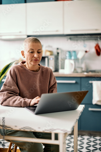 Attractive senior businesswoman dressed casually sitting in a kitchen and working on laptop.