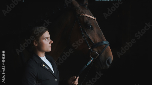 Horsewoman posing with her seal brown horse holding its bridle. The girl is dressed in a black coat. The horse is wearing head jewelry. Grooming horses. Bonding between horse and its master.