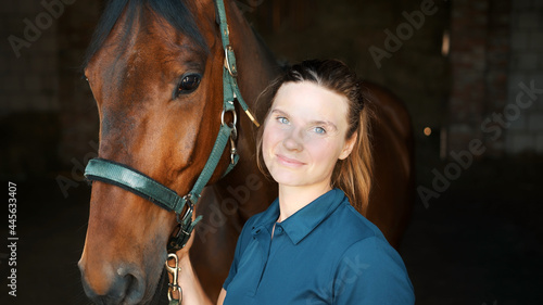 Horse Woman Posing With Her Seal Brown Horse In The Stable Holding The Stallion. Girl smiling at the camera. Holding the bridle of her horse. Horse riding for leisure. 