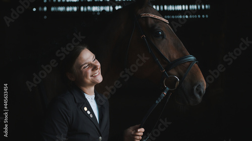 Horsewoman posing with her seal brown horse. Smiling and holding its bridle. The girl is dressed in a black coat. The horse is wearing head jewelry. Grooming horse. Bonding between horse and its