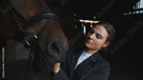 Competitive horsewoman fixing her seal brown horse bridle in the stable. The girl is dressed in a black coat. Preparing her horse for the horse riding competition. 