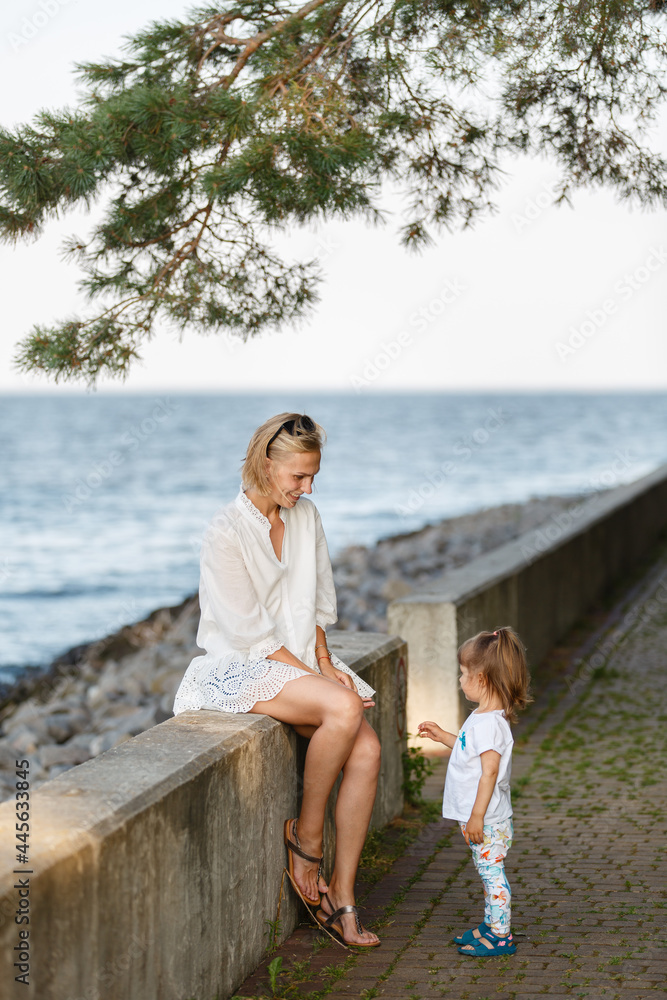 Young woman and toddler girl on sea vacation. Family sits on a beach at sunny day, enjoying summer holidays. Summer lifestyle portrait of pretty family: mom and daughter. Travel local tourism