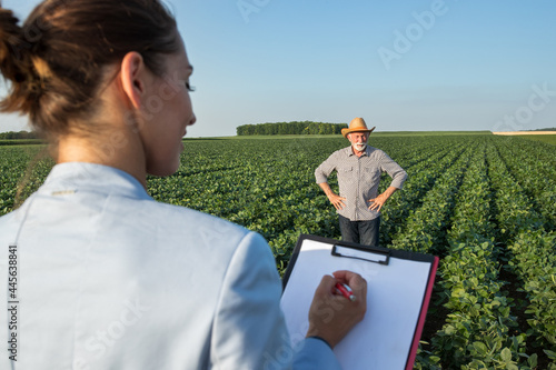 Young female agronomist taking notes uing clipboard with old farmer standing in field in background. photo