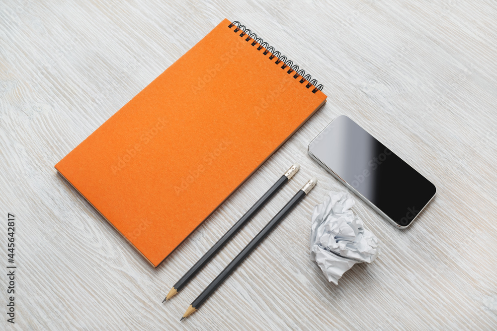 Blank business stationery set on light wooden background. Smartphone, blank orange notepad, pencils and crumpled paper. Blank business stationery set on light wooden background. Smartphone, blank orange notepad, pencils and crumpled paper.