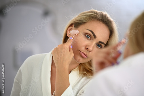 Close-up face portrait of a blonde beautiful woman using jade roller stone massager to perform lymphatic drainage facial massage