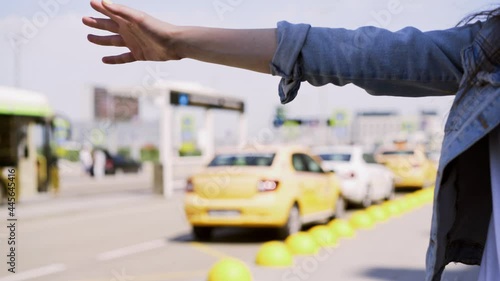 Young woman in blue denim jacket waves hand stopping taxi car standing near grey asphalt road by airport terminal against blurred automobiles close view. Taxi hailing gesture