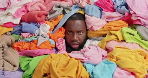 Annoyed dark skinned bearded man shouts loudly overwhelmed by domestic work covered with unfolded clothes squints face fed up of daily chores collects laundry for washing. Housekeeping concept photo