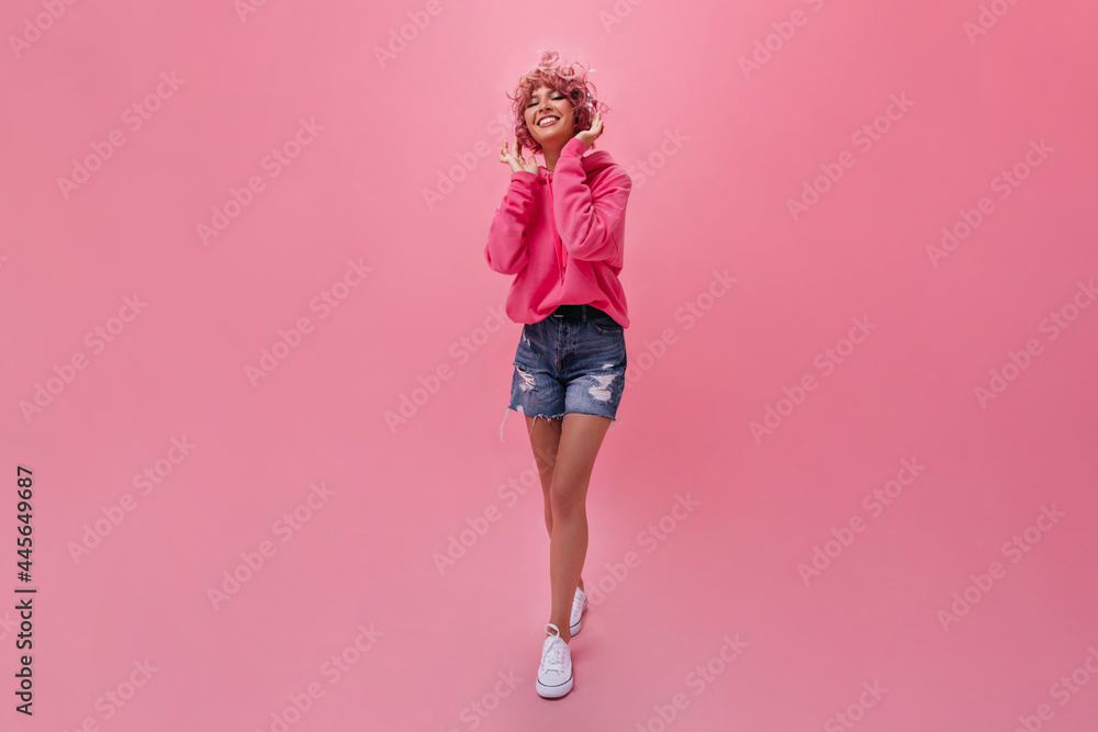 Full-length portrait of pink-haired woman in fuchsia hoodie and denim pants. Cool girl listening to music in headphones and smiling on isolated . Full-length portrait of pink-haired woman in fuchsia hoodie and denim pants. Cool girl listening to music in headphones and smiling on isolated .