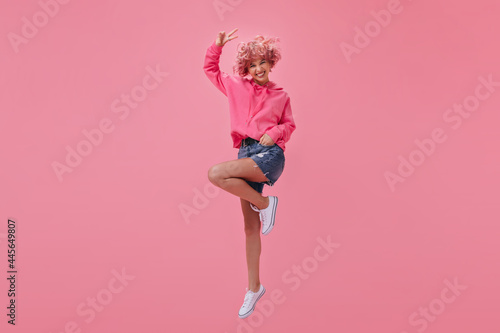 Cool short-haired girl in pink hoodie and denim shorts jumping and smiling. Happy woman with pink hair shows peace sign on isolated.