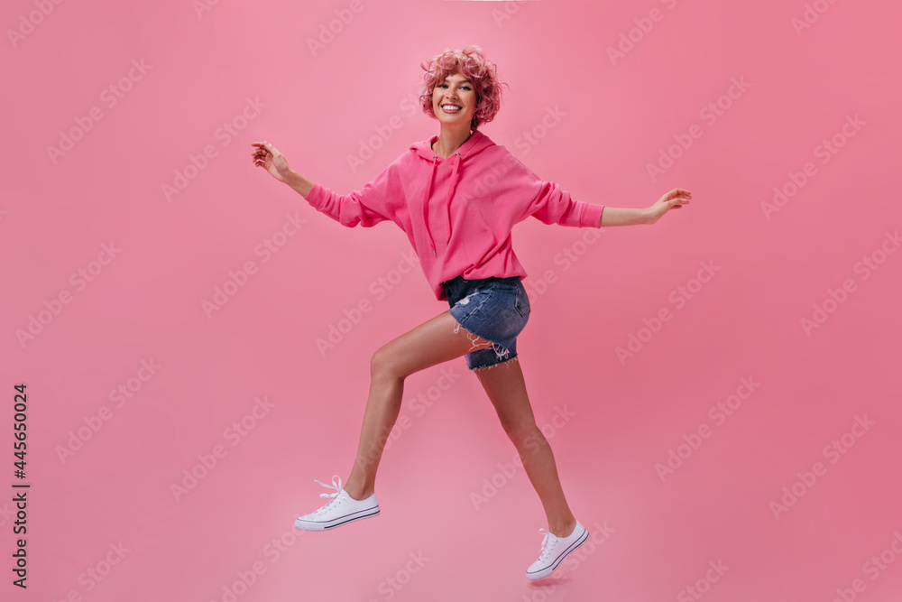 Curly happy woman in denim shorts and hoodie smiles widely and moves on pink background. Pink-haired girl walks on isolated. Curly happy woman in denim shorts and hoodie smiles widely and moves on pink background. Pink-haired girl walks on isolated.