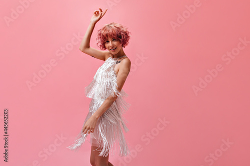 Thin active young curly woman in silver shiny dress moves and spins on isolated. Short-haired girl in festive outfit dances on pink background.