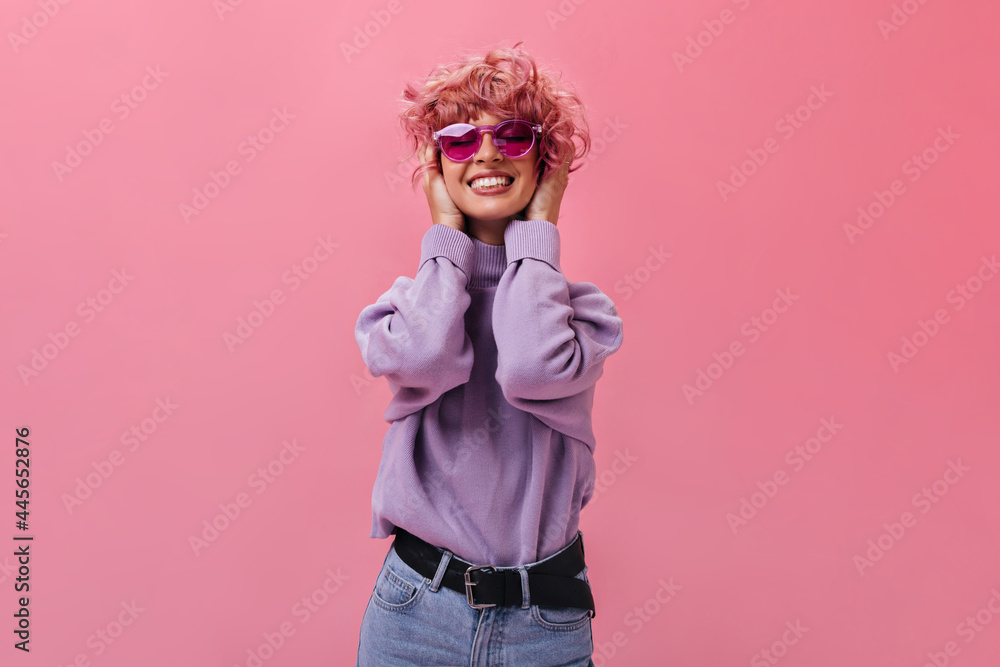 Charming young woman in purple sweater and jeans smiles sincerely. Pink-haired girl in fuchsia sunglasses poses on isolated background . Charming young woman in purple sweater and jeans smiles sincerely. Pink-haired girl in fuchsia sunglasses poses on isolated background .