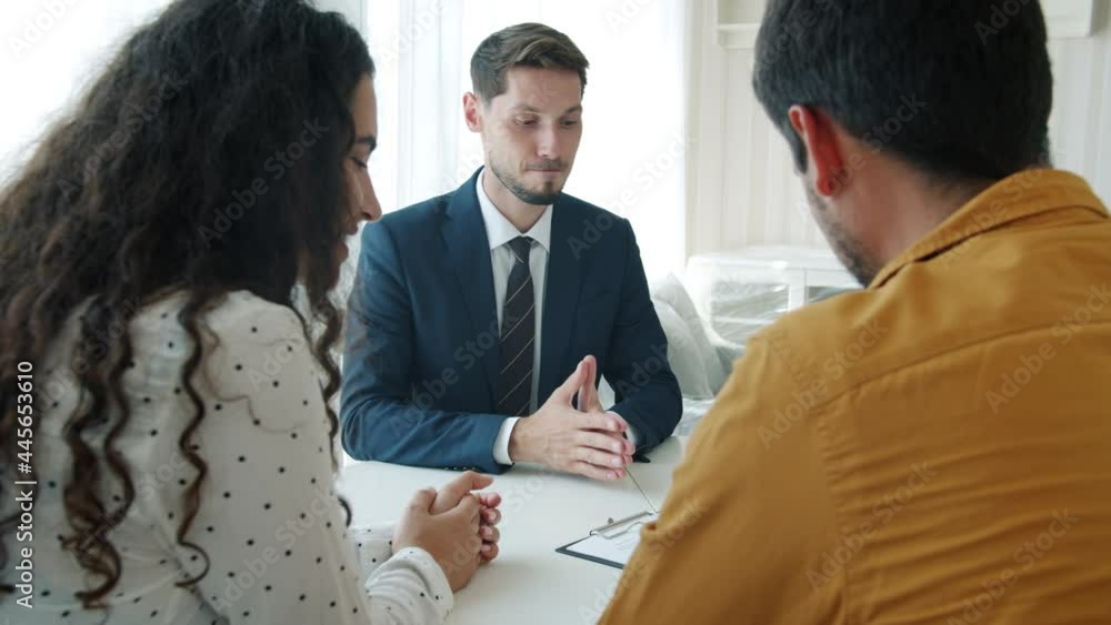 Young couple man and woman are buying house signing agreement hugging talking to happy broker indoors at table. Purchase and sales contract concept.