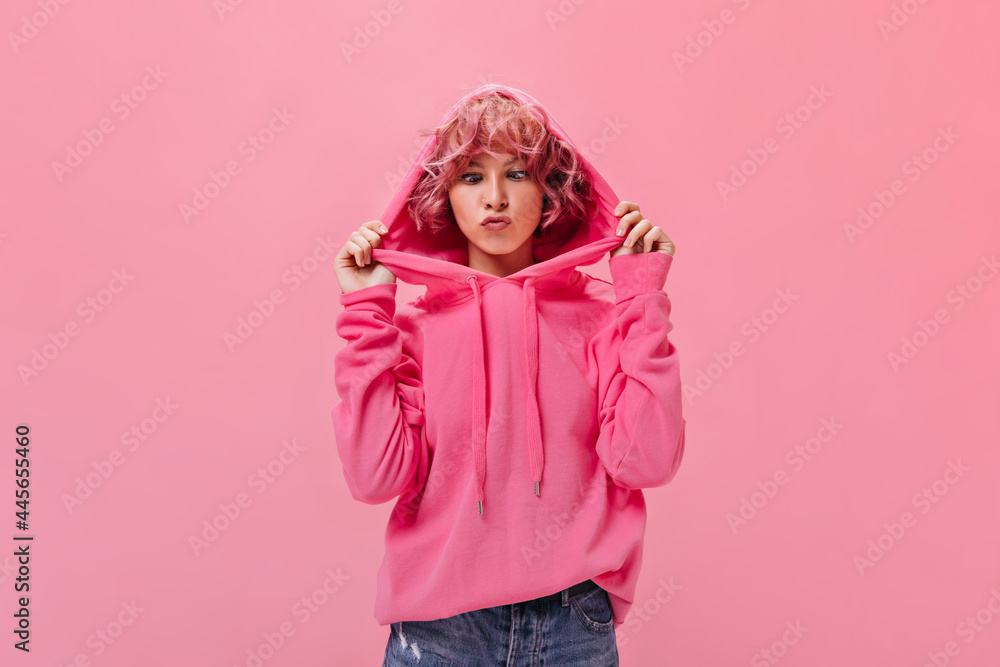 Young good-humored woman in pink hoodie makes funny face. Curly cheerful girl in denim shorts poses on isolated background. Young good-humored woman in pink hoodie makes funny face. Curly cheerful girl in denim shorts poses on isolated background.