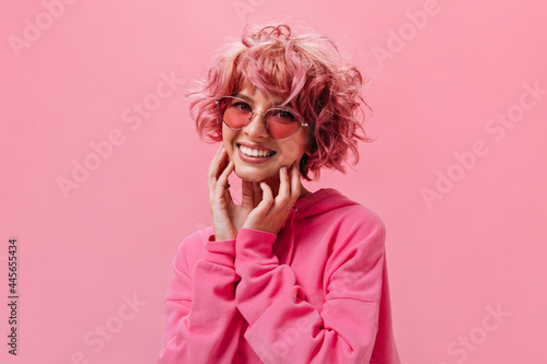 Cheerful happy woman in pink sunglasses poses on isolated background. Portrait of joyful girl in hoodie on pink backdrop.