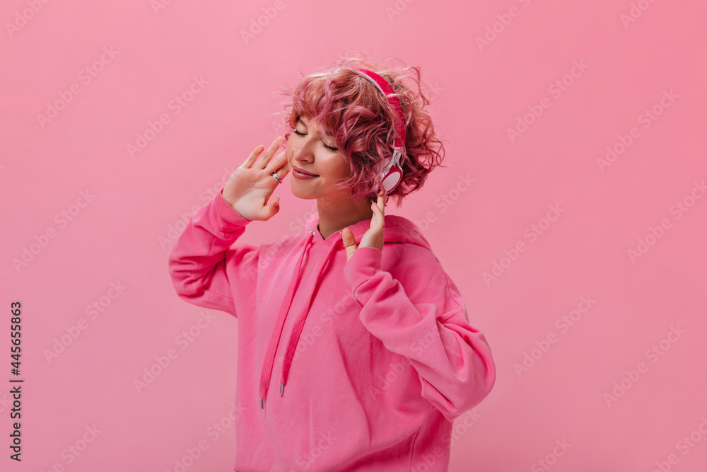 Happy young curly girl in pink hoodie dance and listens to music in headphones on isolated. Short-haired girl smiles sincerely on pink background.