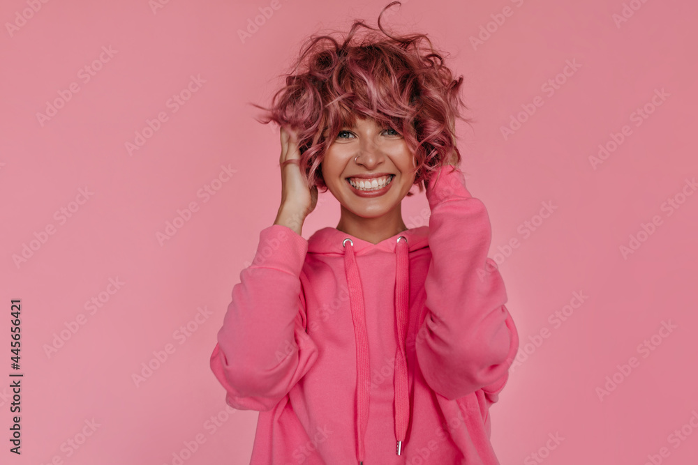 Good-humored curly girl ruffles her pink hair on isolated. Happy pretty tanned woman in hoodie smiles on pink background. Good-humored curly girl ruffles her pink hair on isolated. Happy pretty tanned woman in hoodie smiles on pink background.
