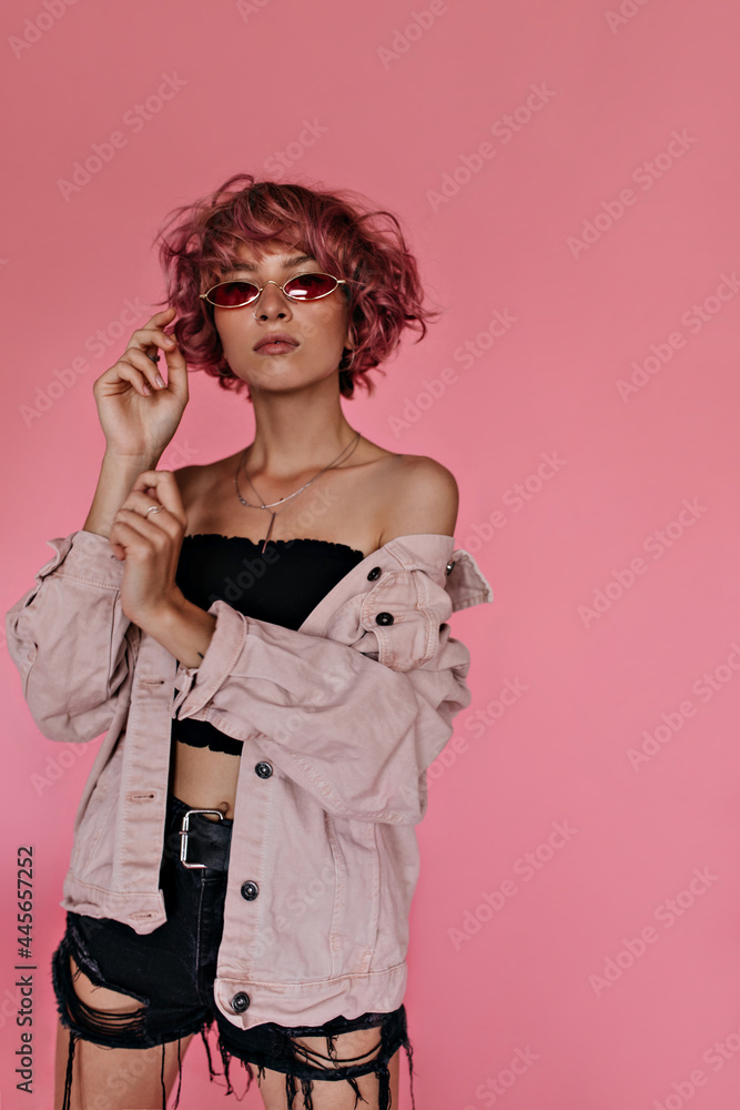 Serious curly woman in black shorts and cropped tee poses on isolated. Portrait of pink-haired girl with tattoos wearing red sunglasses and light denim jacket. Serious curly woman in black shorts and cropped tee poses on isolated. Portrait of pink-haired girl with tattoos wearing red sunglasses and light denim jacket.