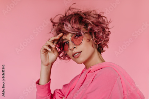 Attractive young woman in pink hoodie and sunglasses looks into camera and touches hair. Pretty girl smiles on isolated background.