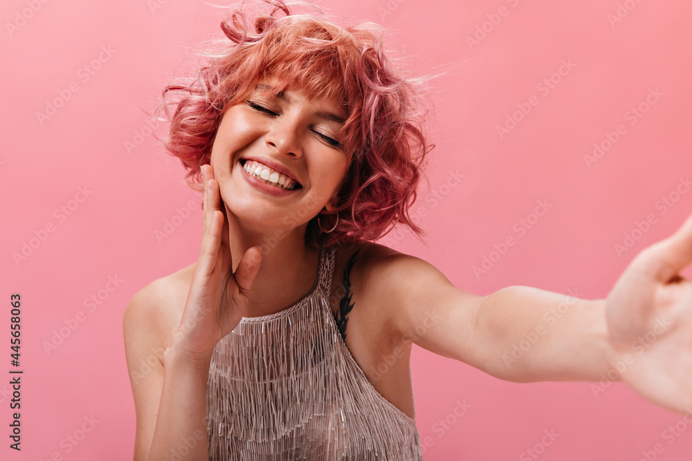 Cheerful curly woman in shiny top takes selfie. Happy joyful girl in silver dress smiles on isolated pink background. Cheerful curly woman in shiny top takes selfie. Happy joyful girl in silver dress smiles on isolated pink background.