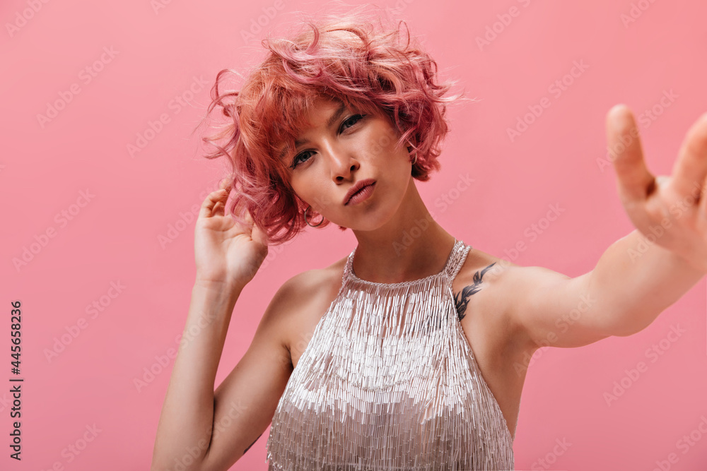 Close-up inside portrait of attractive tanned curly woman with tattoos dressed in silver shiny festive outfit taking selfie on pink isolated background . Close-up inside portrait of attractive tanned curly woman with tattoos dressed in silver shiny festive outfit taking selfie on pink isolated background .