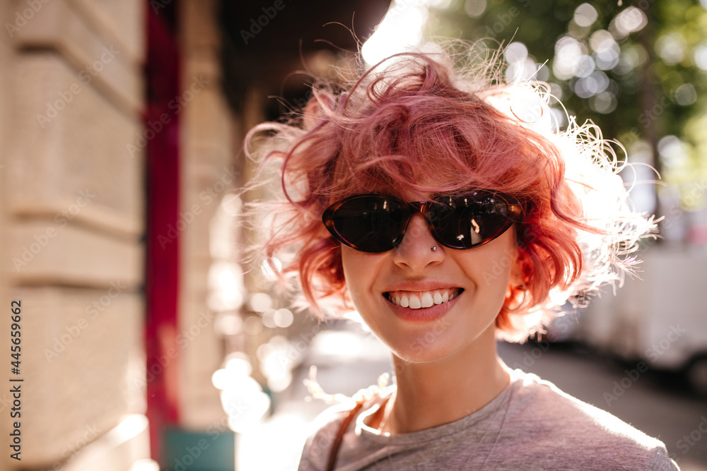 Curly lady in sunglasses smiles widely outside. Close-up portrait of happy pink-haired woman in grey tee laughing and walking on street. Curly lady in sunglasses smiles widely outside. Close-up portrait of happy pink-haired woman in grey tee laughing and walking on street.