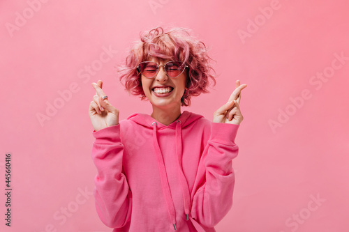 Cool young woman in pink sunglasses and bright hoodie smiles widely with closed eyes and crosses fingers on isolated background .