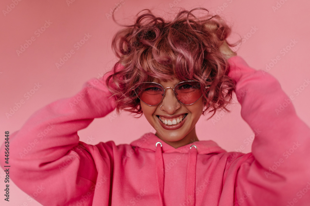 Curly cute woman in pink hoodie ruffles hair and smiles widely. Charming girl in sunglasses poses on isolated background. Curly cute woman in pink hoodie ruffles hair and smiles widely. Charming girl in sunglasses poses on isolated background.