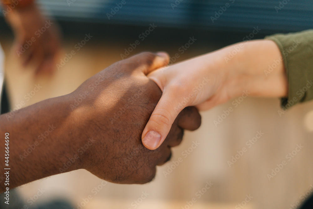 Fototapeta premium Close-up top view of handshake of Caucasian woman and black male indoors at home office, selective focus. Concept of handshaking black and white hand, interracial friendship and cooperation.