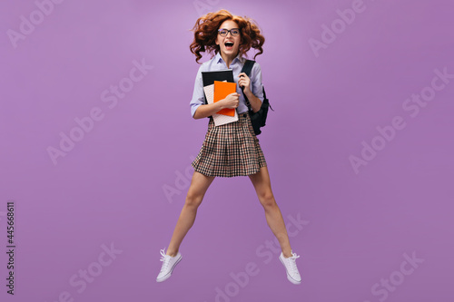 Joyful young woman in blue blouse and round eyeglasses jumps on purple background and holds notebooks and black backpack..