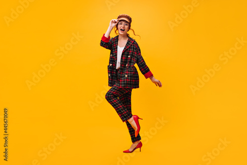 Happy woman in plaid outfit moves on orange background. Cheerful girl in brown cap and eyeglasses has fan on isolated backdrop..