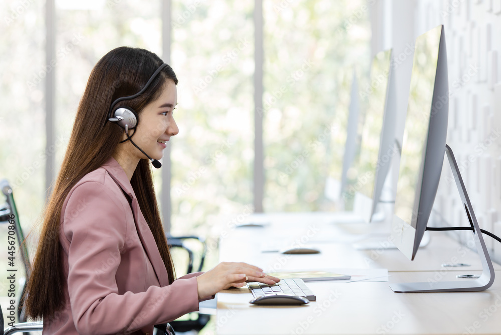 Obraz premium Asian young beautiful happy confident professional female operator wears pink formal suit and microphone headset sitting talking solving problem with customer while typing information with keyboard