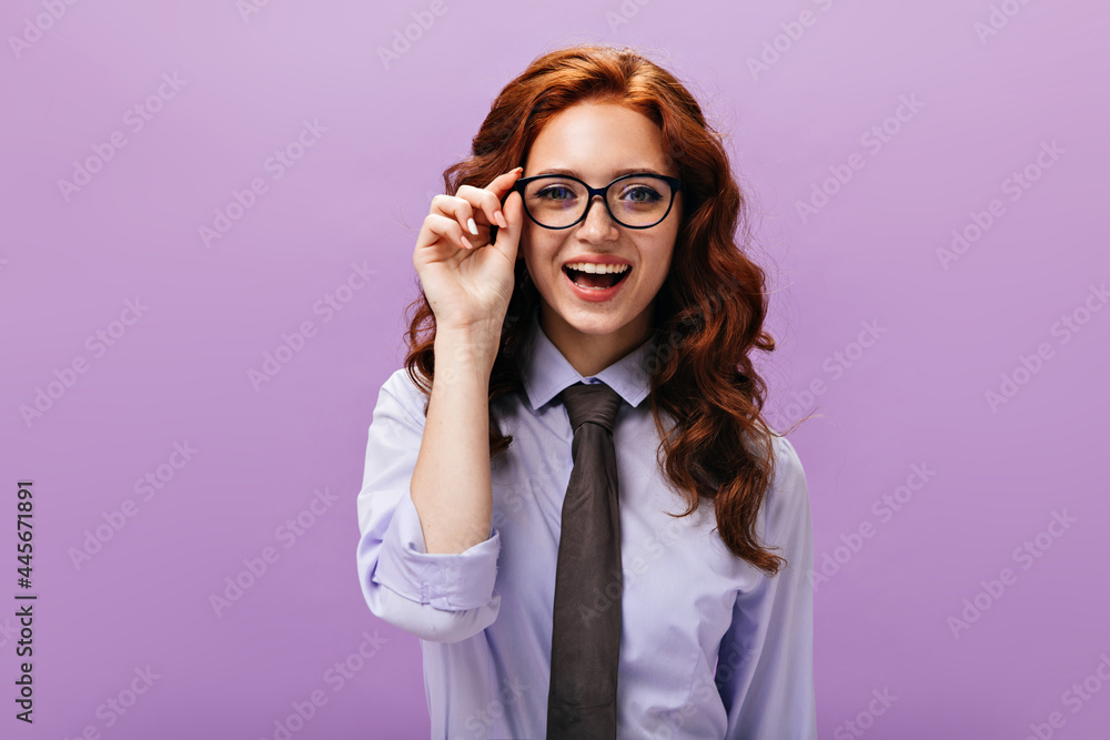 Ginger girl in eyeglasses looks into camera on purple background. Joyful young woman with wavy hair in lilac shirt and black tie posing.. Ginger girl in eyeglasses looks into camera on purple background. Joyful young woman with wavy hair in lilac shirt and black tie posing..