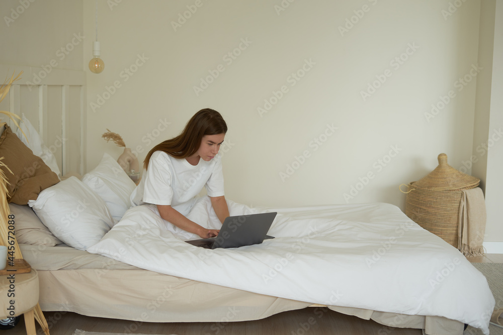 a young girl in a white T-shirt in the morning is on the bed under a blanket working on a computer and talking on the phone enthusiastically
