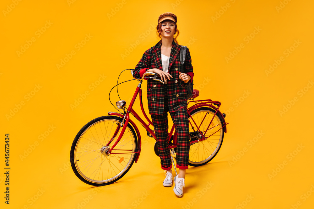 Naklejka premium Woman in plaid suit leans on bicycle. Stylish wonderful girl in cool clothes, white sneakers and brown cap posing on isolated background..