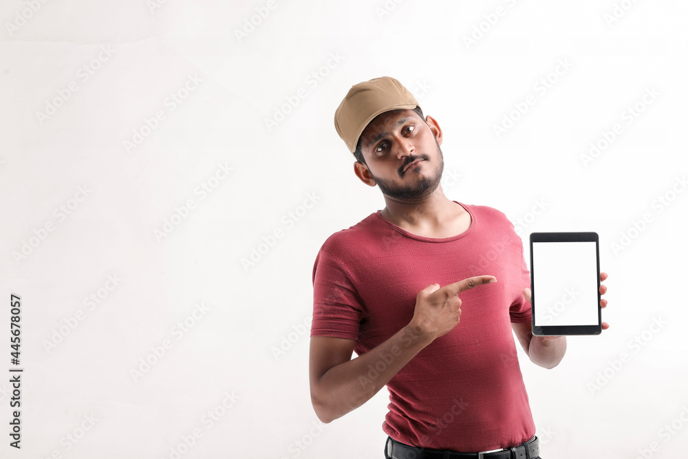 Portrait of a excited happy young delivery man in cap standing over white background. Looking camera showing display of mobile phone.